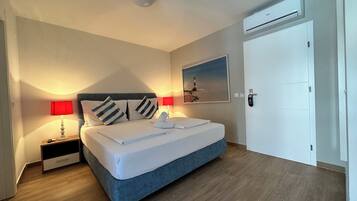 A neatly made bed with a blue headboard, two bedside lamps, a wall-mounted air conditioner, and a door with a handle.