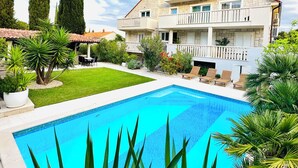 Un edificio de dos pisos con piscina, rodeado de follaje y árboles altos.