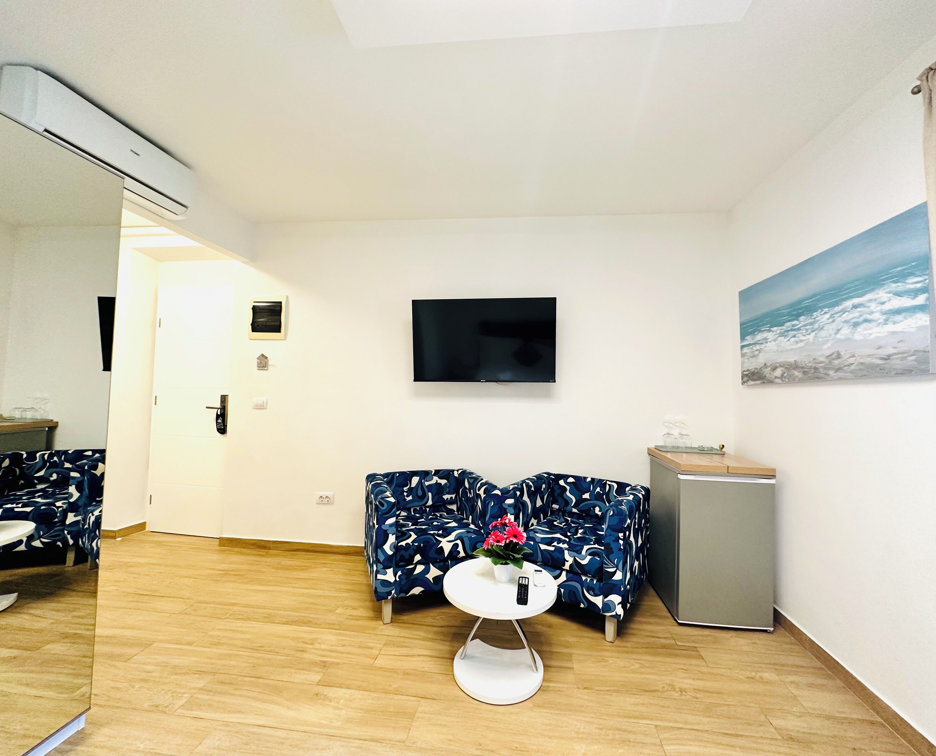 A hotel room with a flat-screen TV, a painting of the ocean, a small table with a vase of flowers, and a sofa with a blue and white pattern.