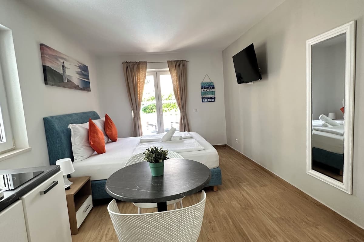 A compact hotel room with a bed, a small dining table, a kitchenette, and a TV mounted on the wall.