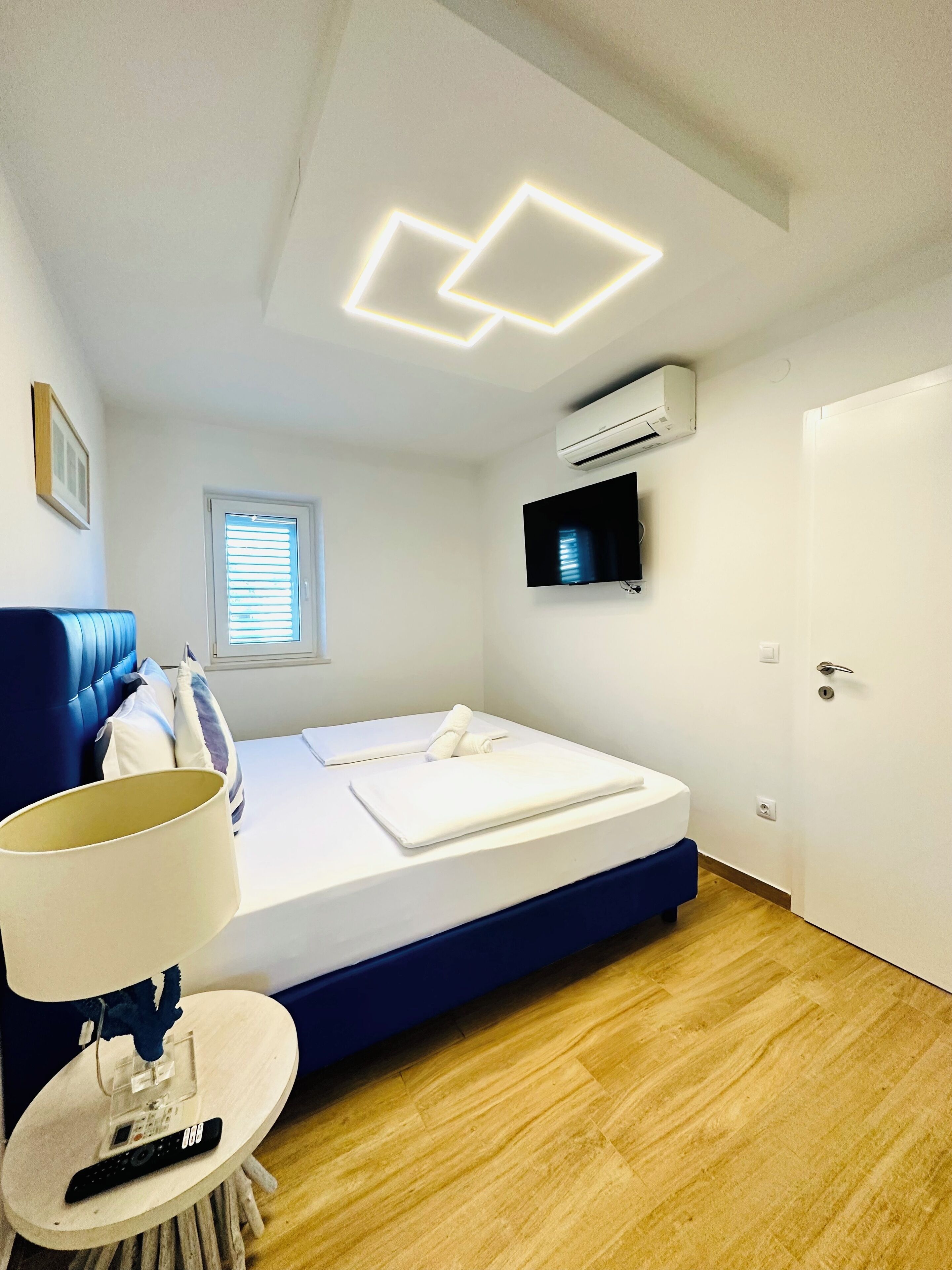 A modern bedroom with a bed, a TV, an air conditioner, and a window with blinds.