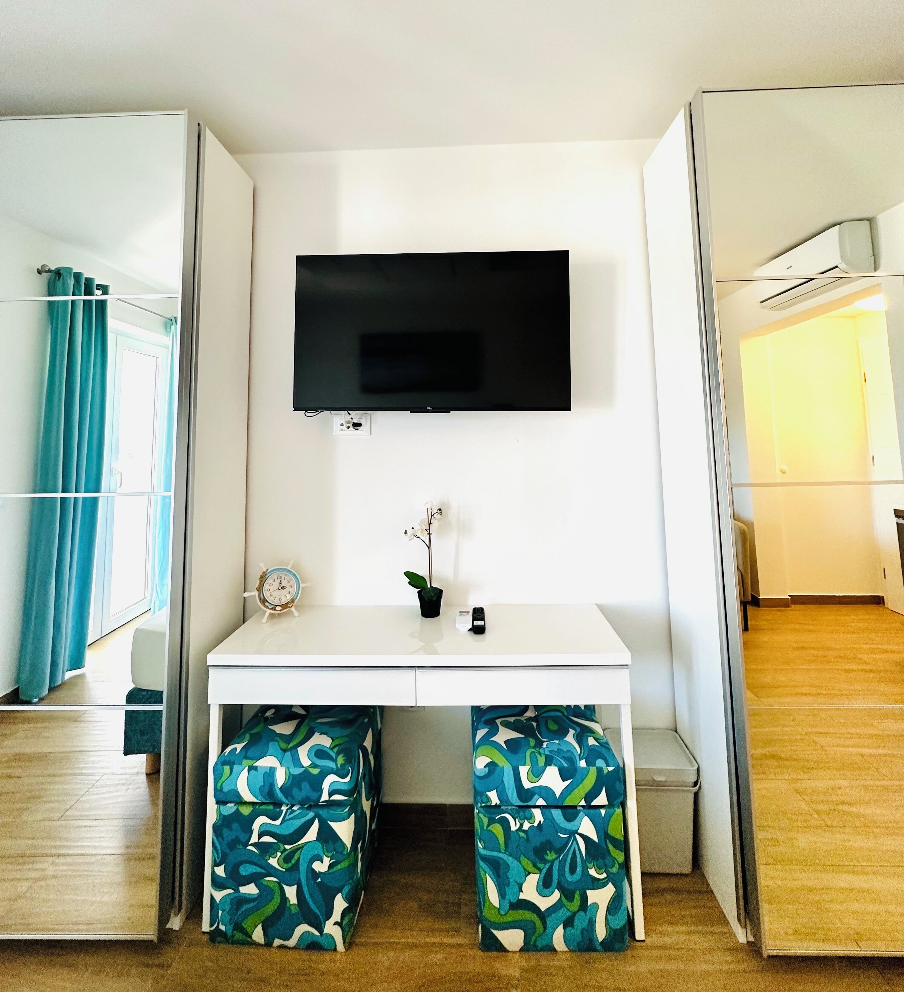 Una habitación compacta con un escritorio blanco, un televisor de pantalla plana montado encima, una pequeña planta en maceta y dos otomanas a juego.