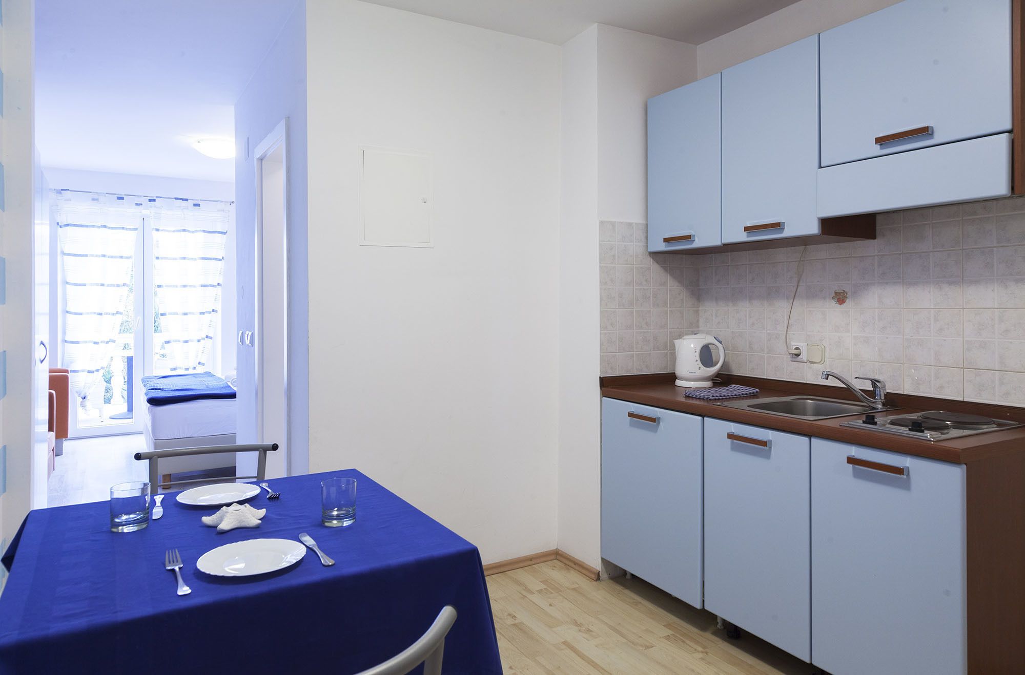 A small kitchen with a sink, a kettle, and cabinets. There is a dining table with plates and cutlery.