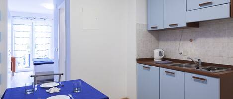 A small kitchen with a sink, a kettle, and cabinets. There is a dining table with plates and cutlery.