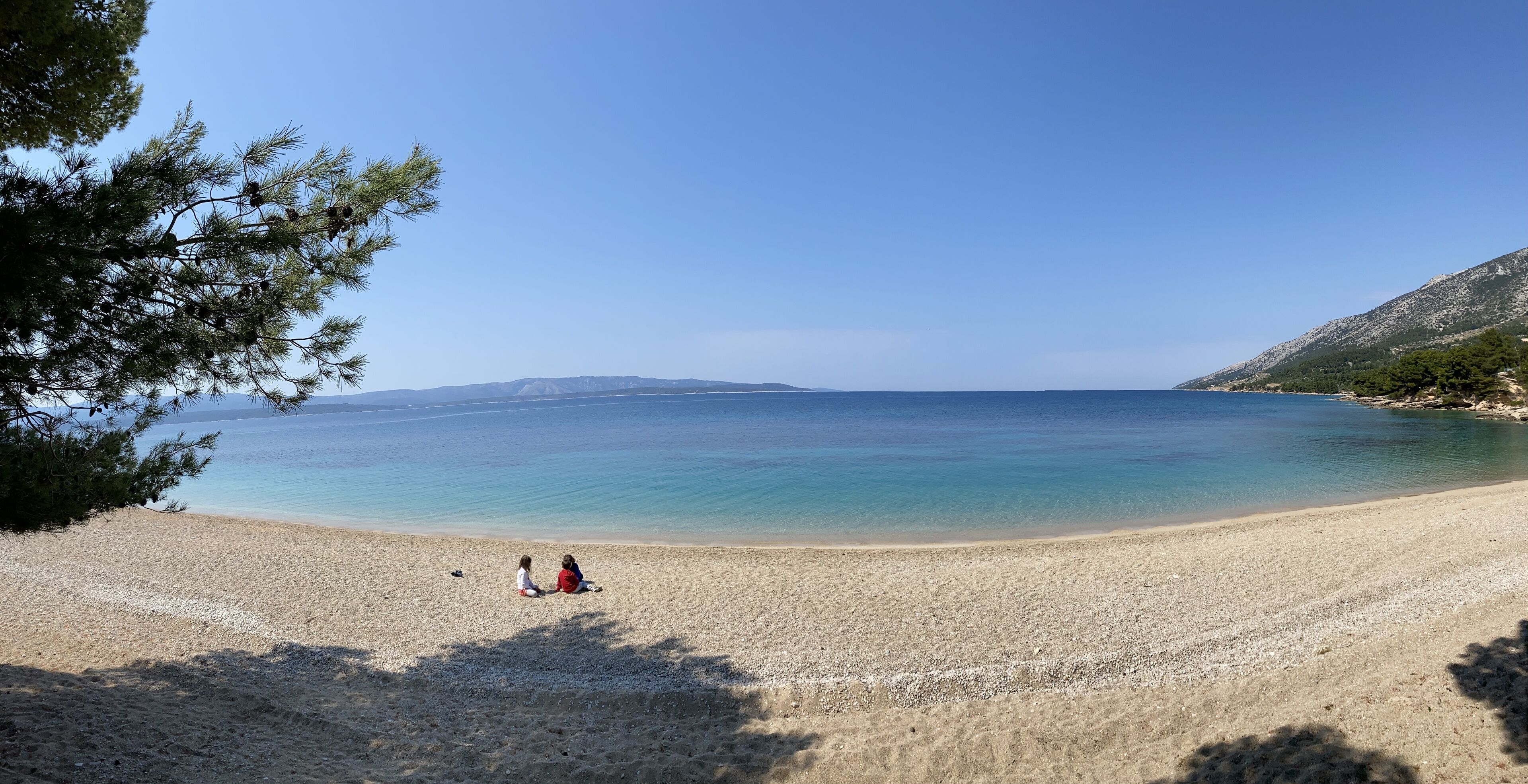 A beach with two people sitting on the sand, a clear sky, and a calm sea.