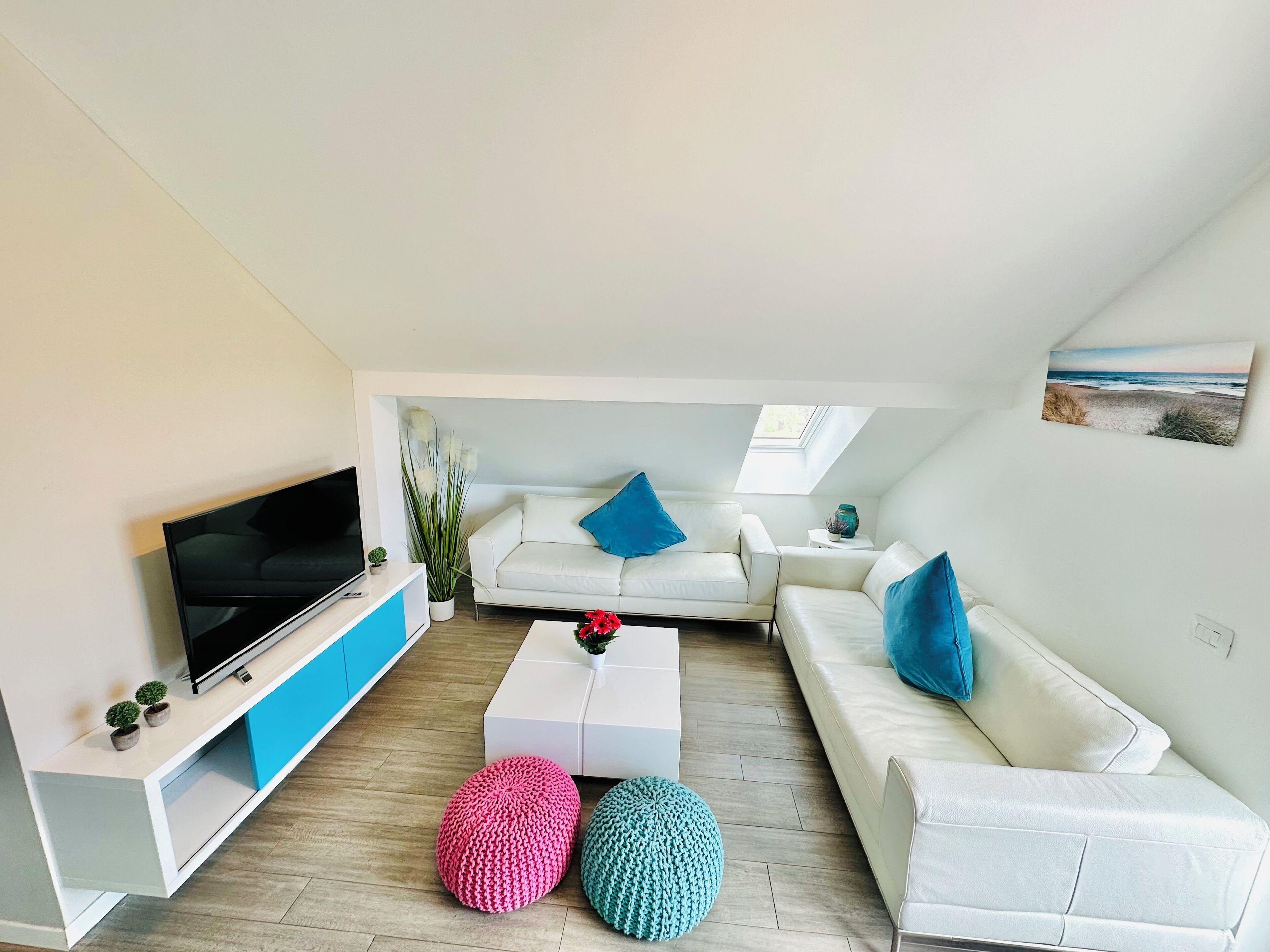 Ein modernes Wohnzimmer mit Fernseher, weißen Sofas und bunten Poufs.