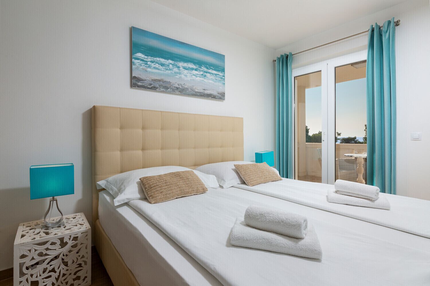 A hotel room with a large bed, two pillows, a headboard, a bedside table with a lamp, and a painting of a beach scene.
