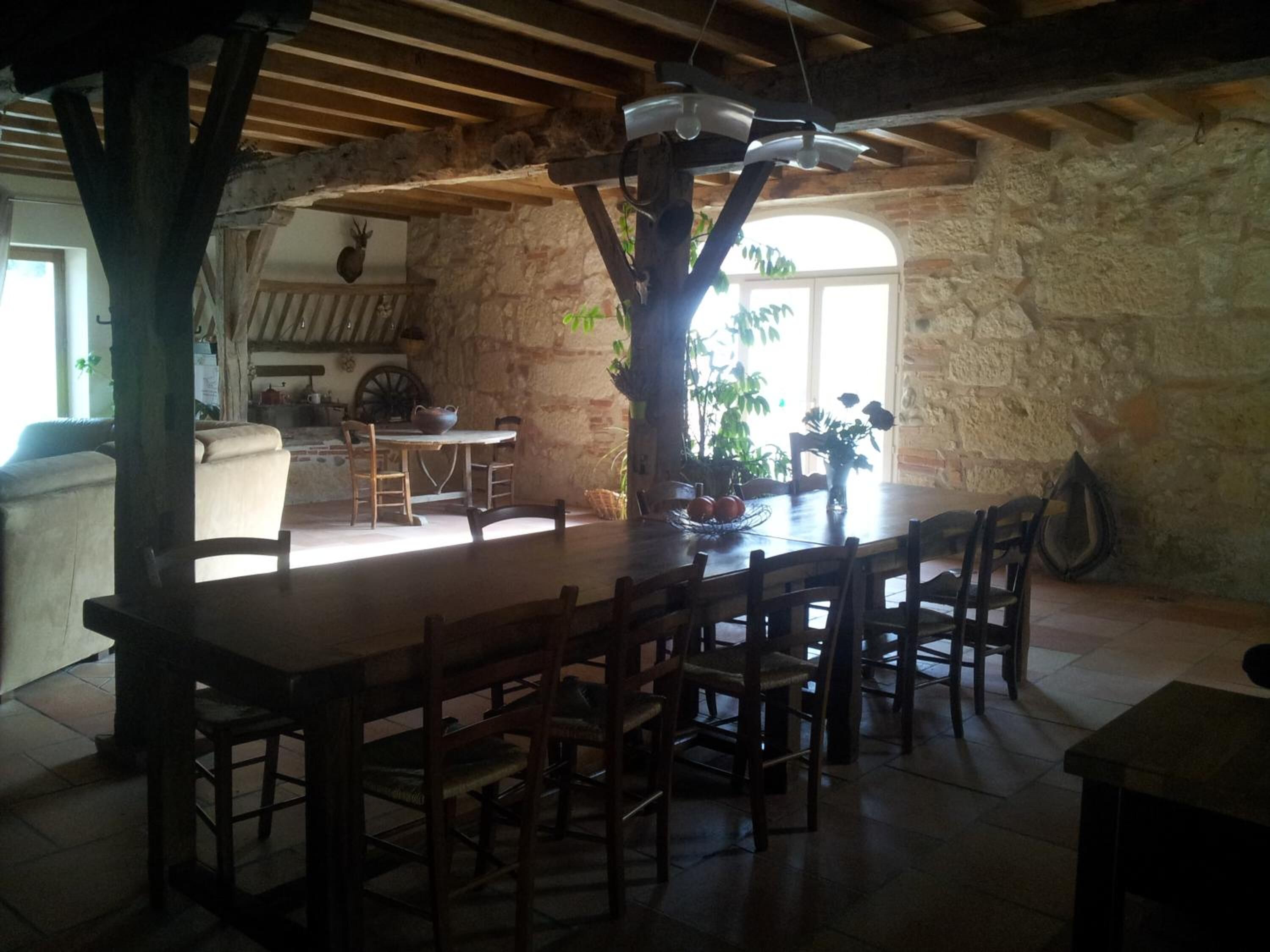 Un comedor rústico con una gran mesa de madera y sillas, un muro de piedra, y vistas a una cocina y zona de estar.