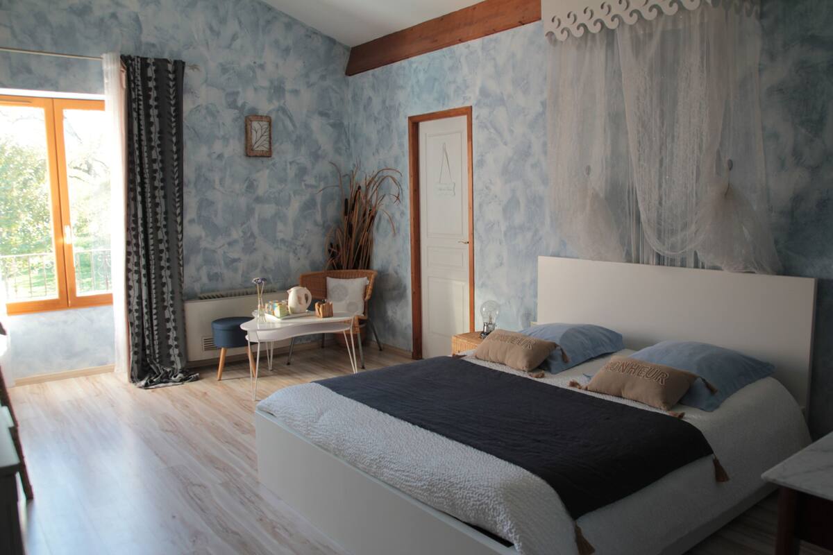 Ein Schlafzimmer mit Bett, Nachttisch, Stuhl, einem Fenster mit Vorhängen und einer Tür.