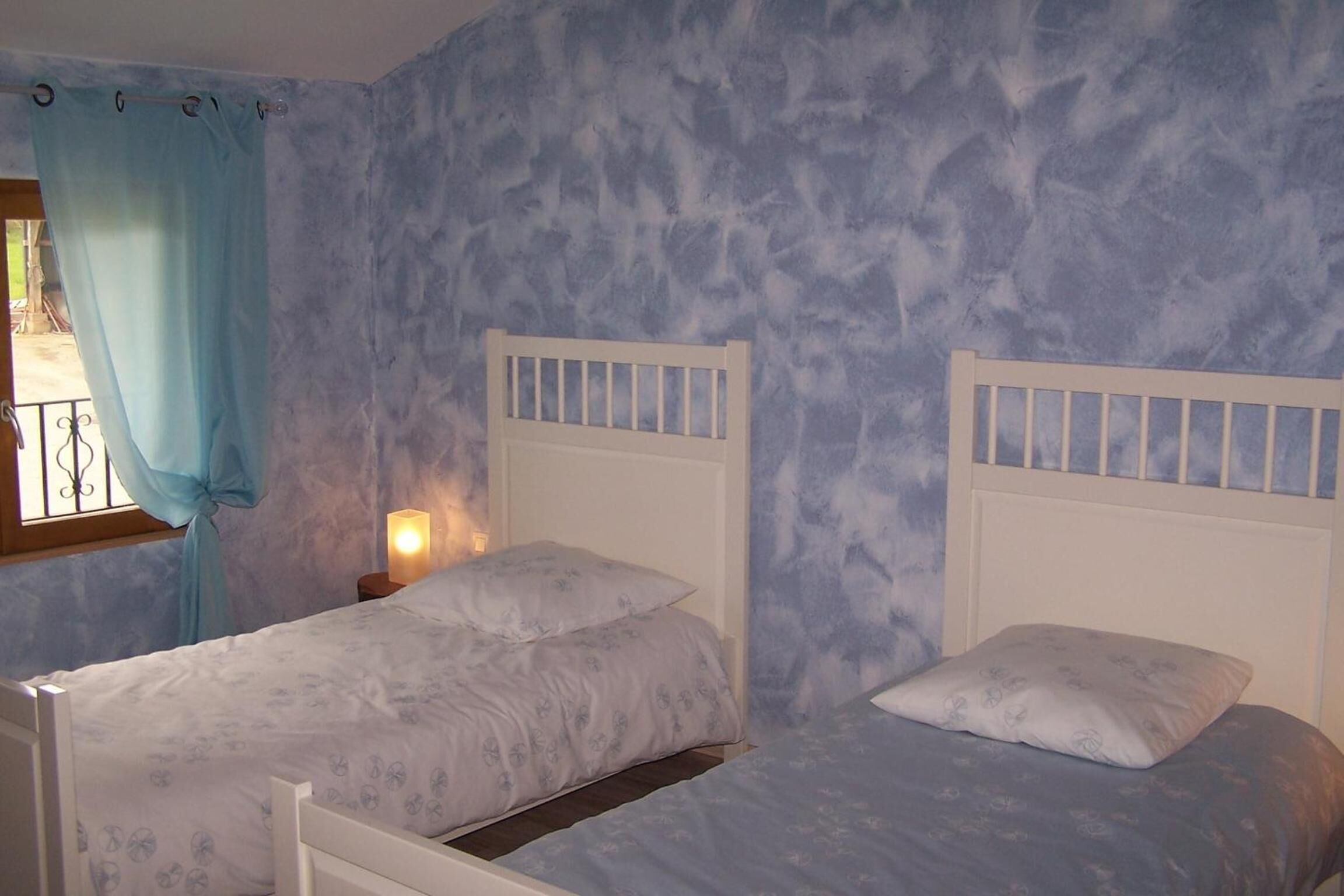 A bedroom with two beds, a window with curtains, and a nightstand with a lamp.