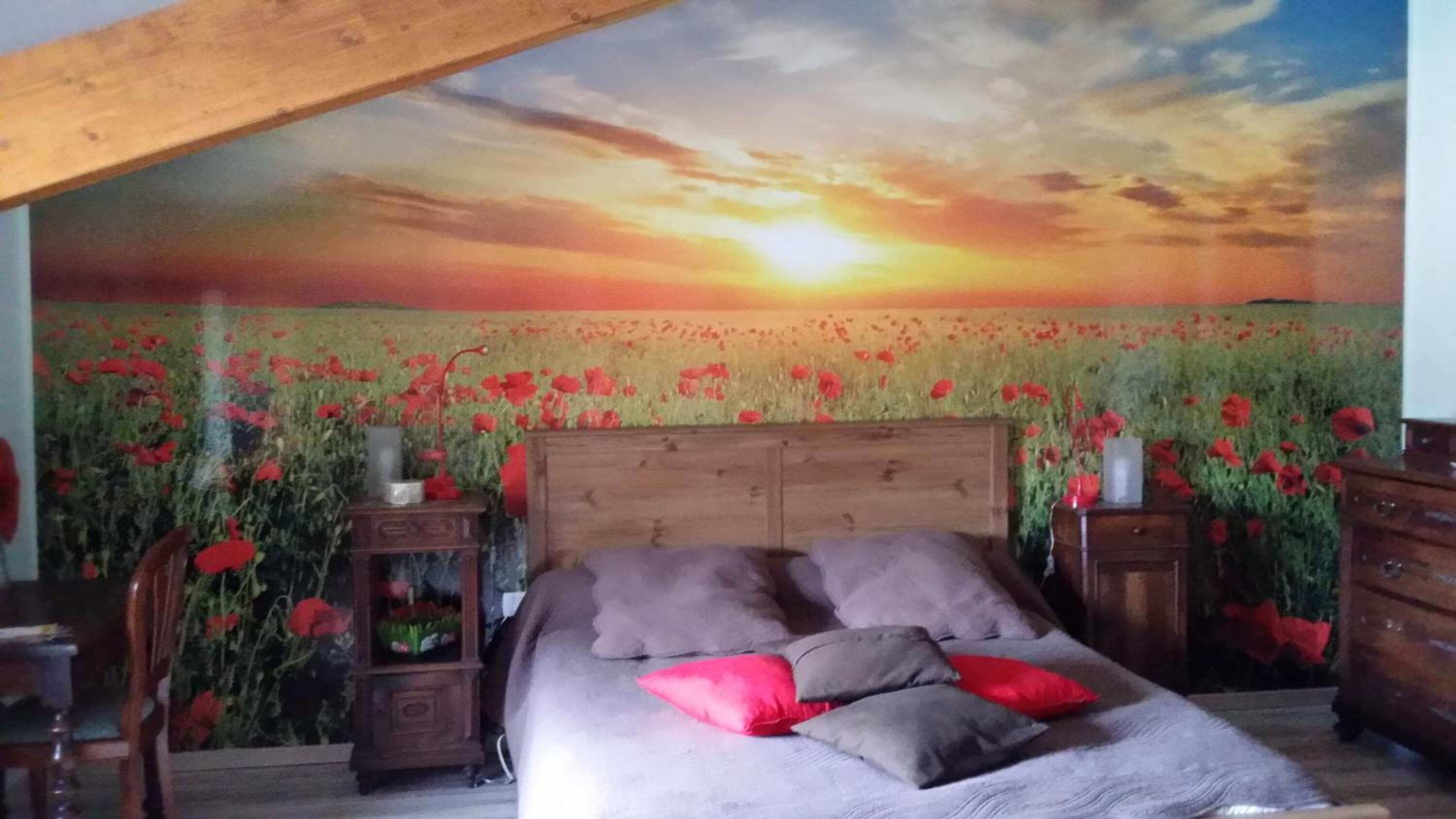 Een slaapkamer met een muurschildering van een zonsondergang boven een veld met klaprozen, een houten hoofdbord en een bed met grijze en rode kussens.
