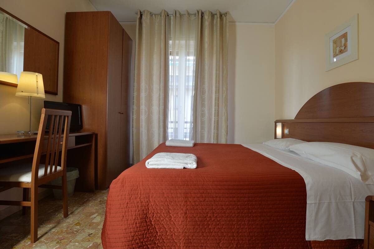 Een hotelkamer met een bed, een bureau, een stoel, een lamp en een raam met gordijnen.