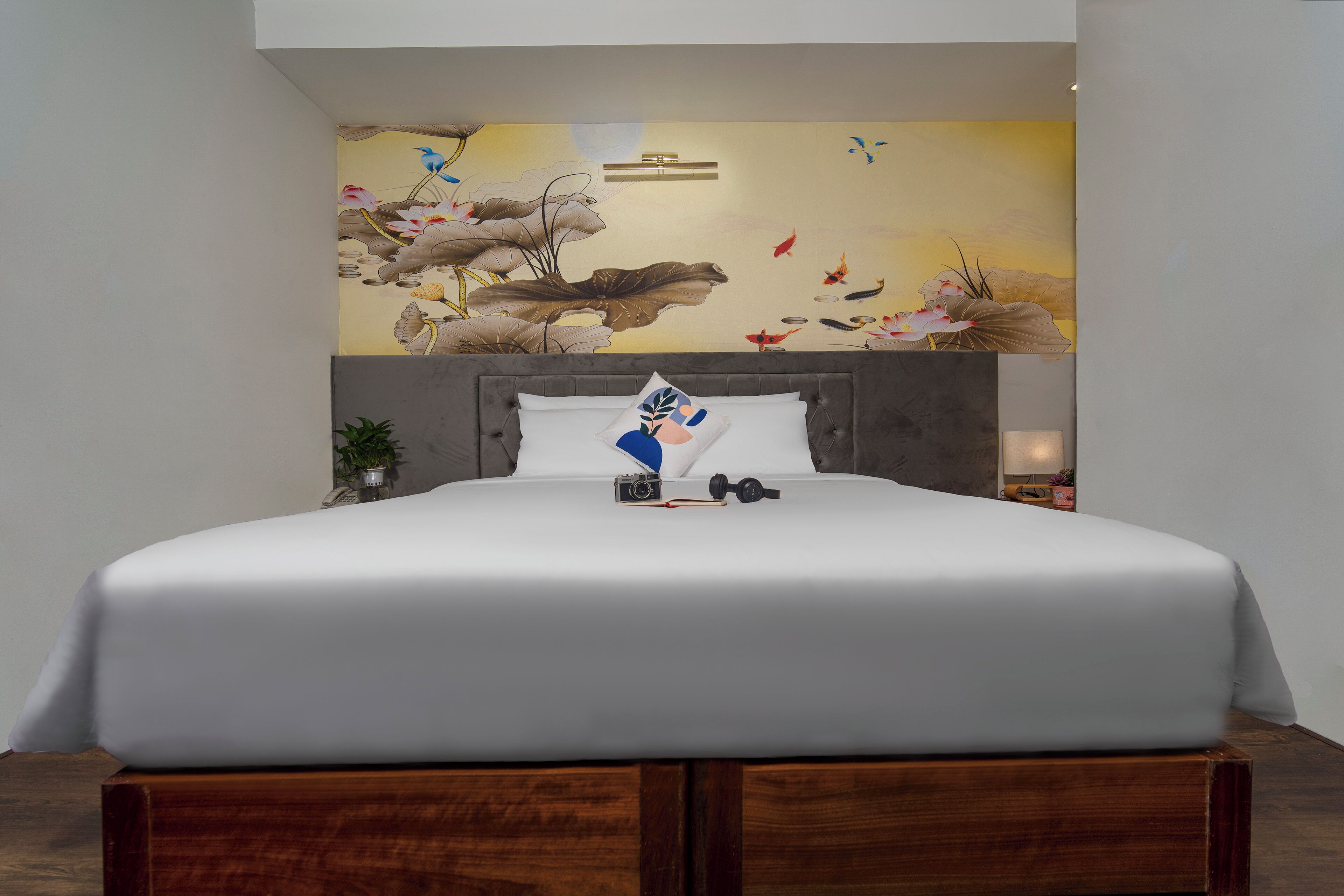 Una cama bien hecha con edredón blanco y almohadas, una mesita de noche con lámpara y un mural en la pared que representa una escena natural.