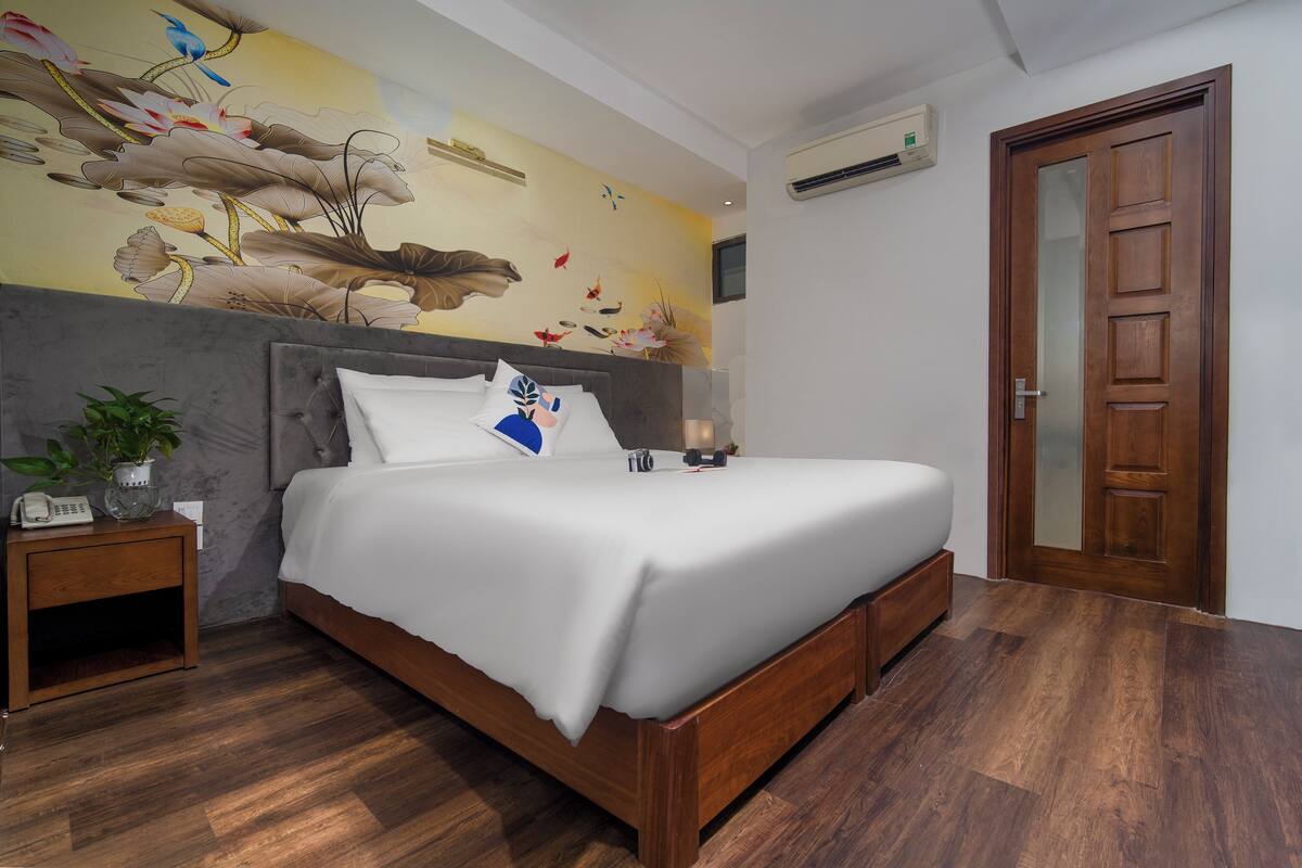 A neatly made bed with white linens, a wooden headboard, a bedside table with a lamp, and a wall mural.