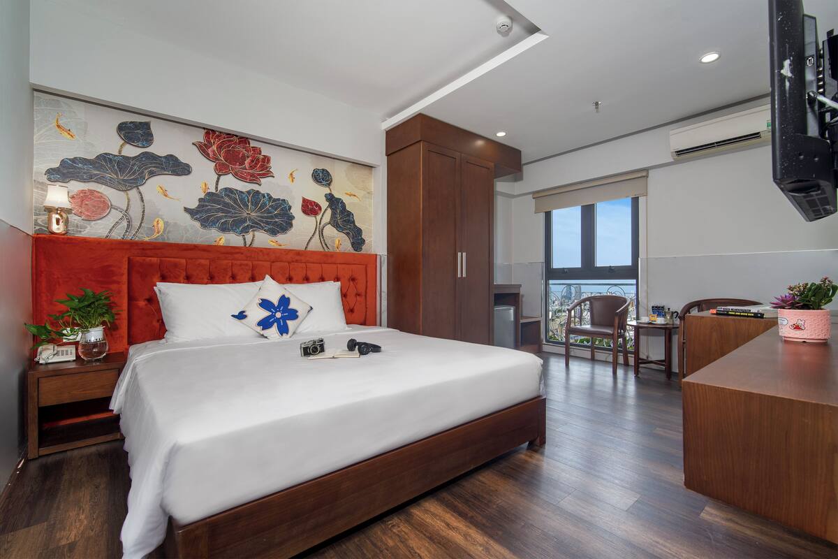A modern bedroom with a large bed, a red headboard, a decorative wall mural, a desk, and a view through a large window.