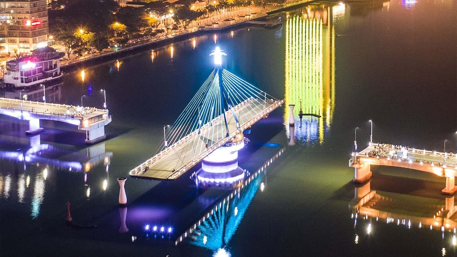A nighttime view of a cable-stayed bridge with illuminated cables and a central tower, spanning over a body of water with a cityscape in the background.