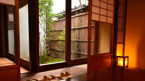 Una habitación japonesa tradicional con suelo de tatami, una puerta corredera que da a un jardín y una mesita con zapatillas.