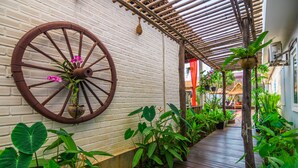 A wooden pathway lined with lush green plants and a large wooden wheel mounted on a white brick wall. - Wheel Garden Residence (Siem Reap)