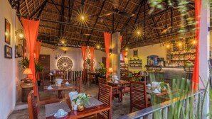 A restaurant with wooden furniture, a thatched roof, and a brick wall with a large circular window. - Wheel Garden Residence (Siem Reap)