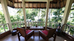 A covered outdoor seating area with two wooden chairs and a table, surrounded by a white railing, overlooking a lush garden with palm trees. - Felicianas Resort (Tejakula)