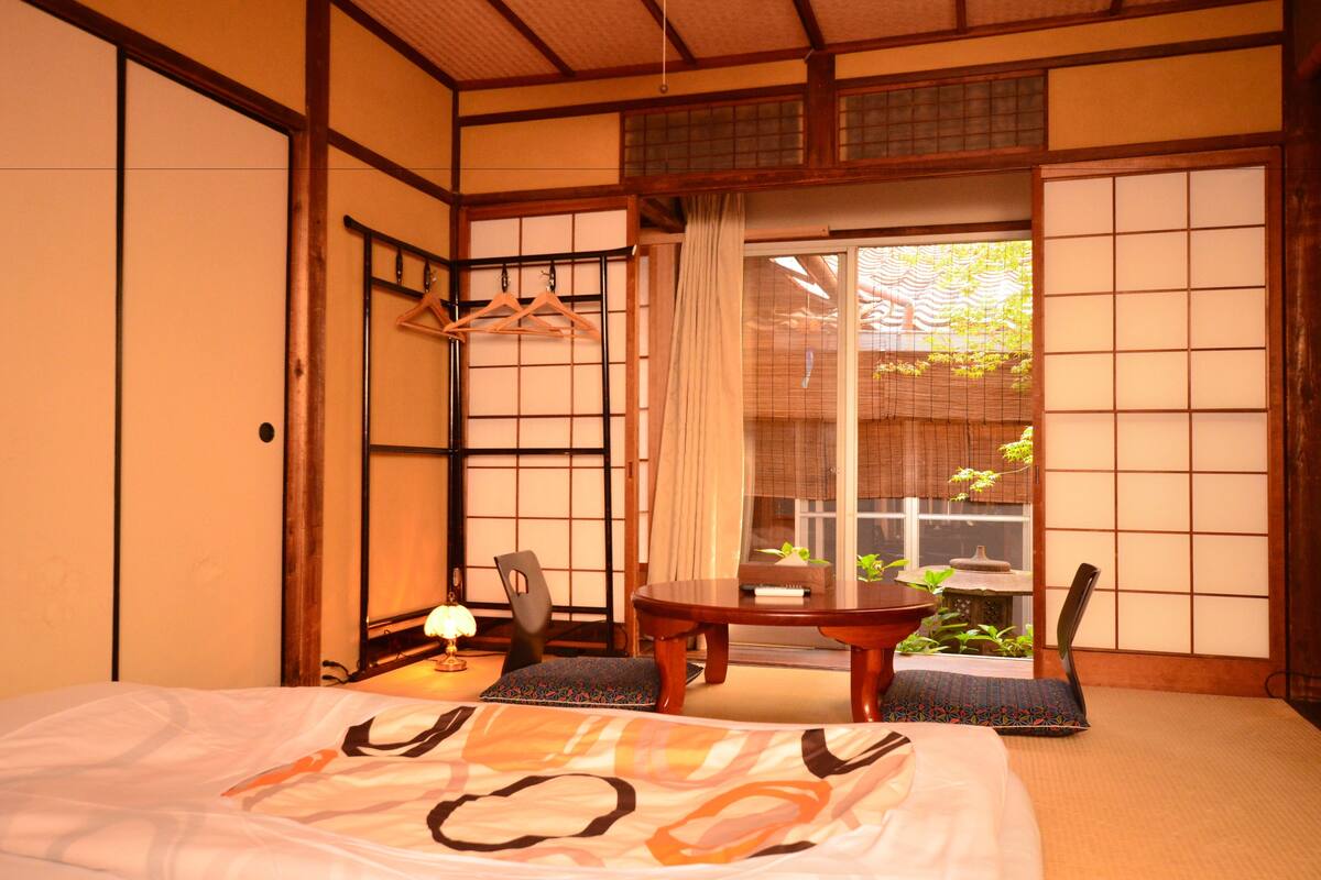 Une pièce de style japonais traditionnel, avec un sol recouvert de tatamis, une petite table basse avec un coussin, un porte-manteau suspendu et une porte coulissante donnant sur un jardin.