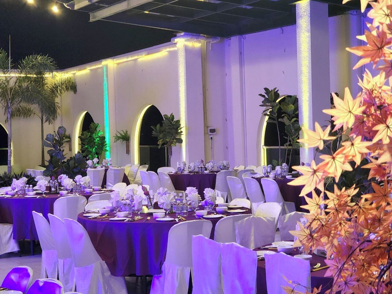 Un salón de banquetes con mesas redondas, sillas blancas y manteles morados.