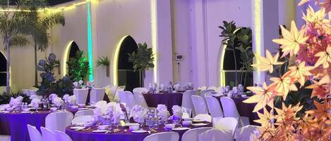 Un salón de banquetes con mesas redondas, sillas blancas y manteles morados.