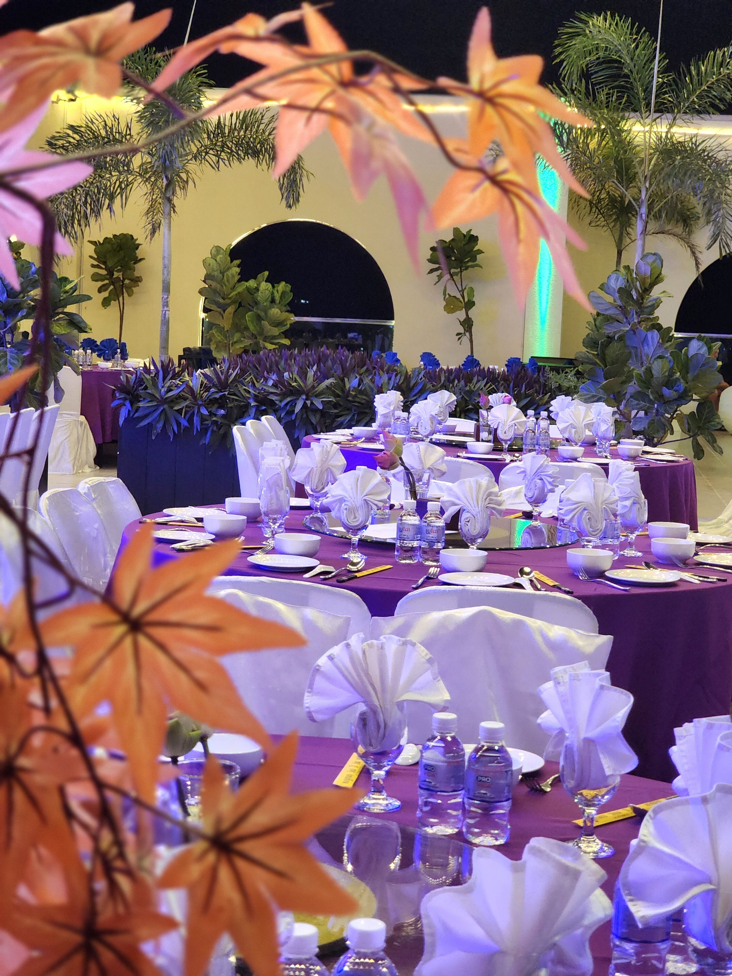 A banquet hall with round tables set for dining, surrounded by lush greenery and decorative plants.