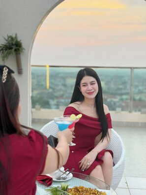 Une femme vêtue d’une robe rouge est assise à une table, un verre à la main, à l’extérieur, avec une vue sur le paysage urbain.