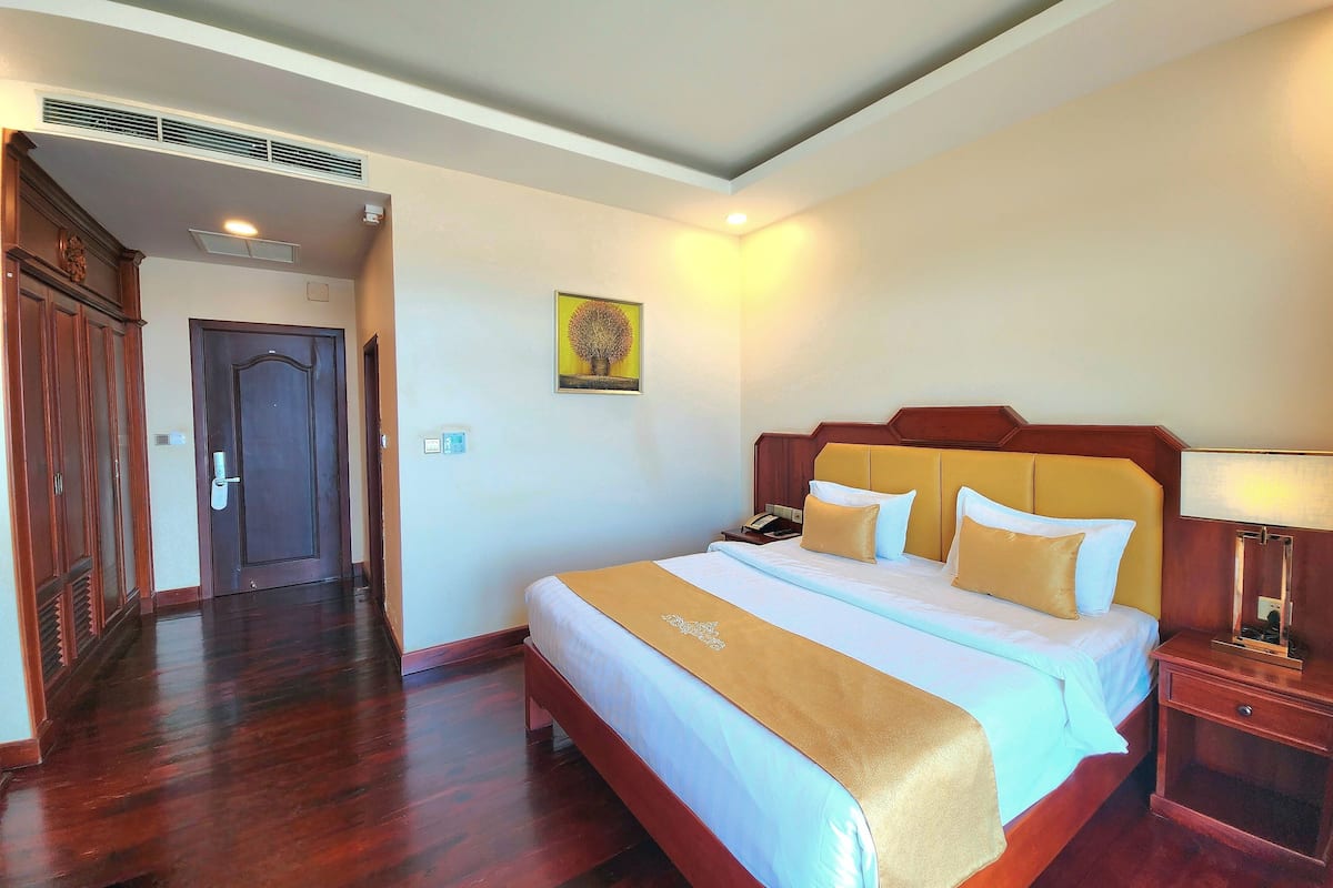 Une chambre d’hôtel avec un lit en bois, une table de chevet, une commode, une lampe fixée au mur, une porte et un couloir.