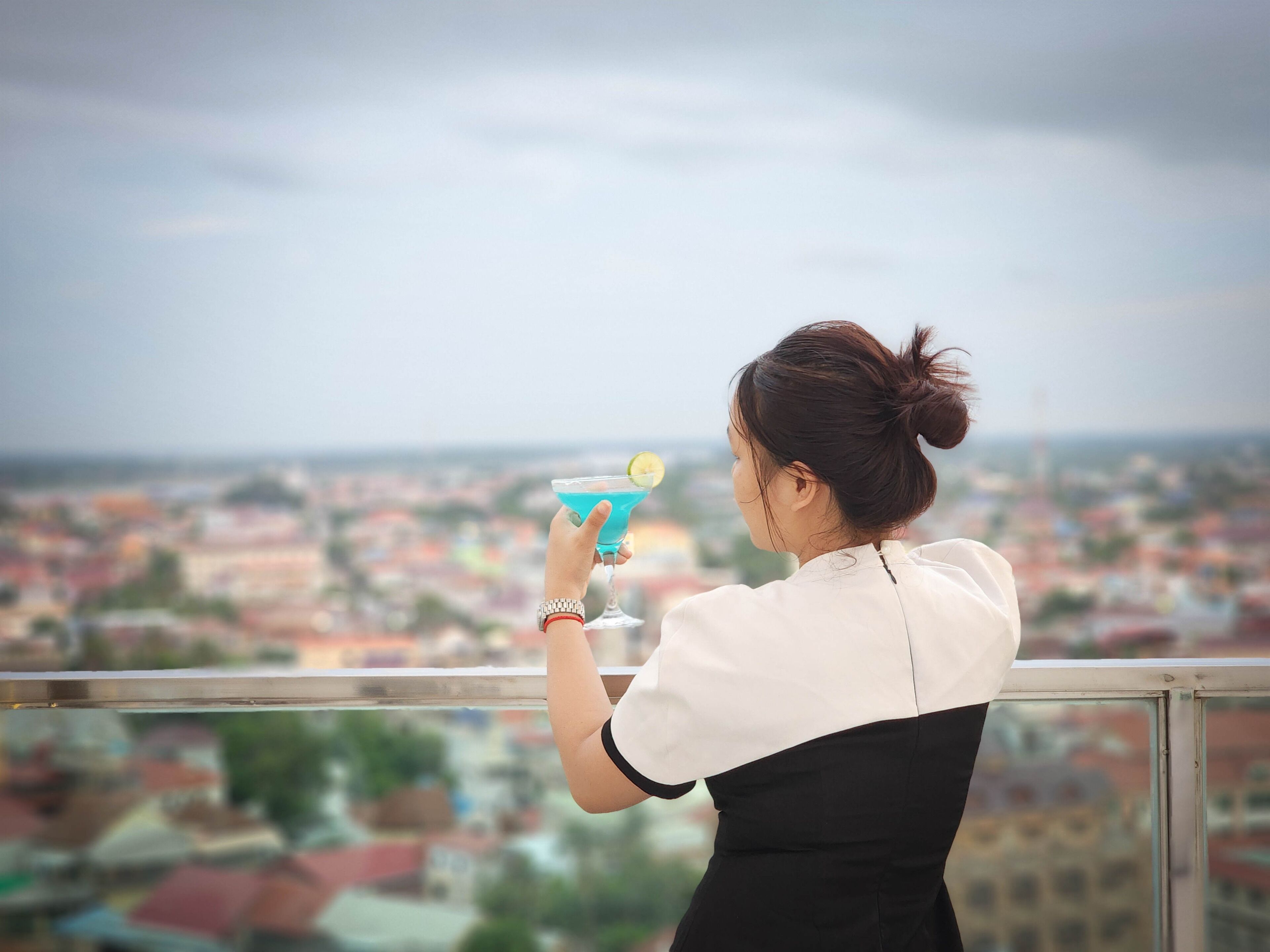 A person holding a blue cocktail glass with a lime wedge, standing on a balcony with a cityscape view.