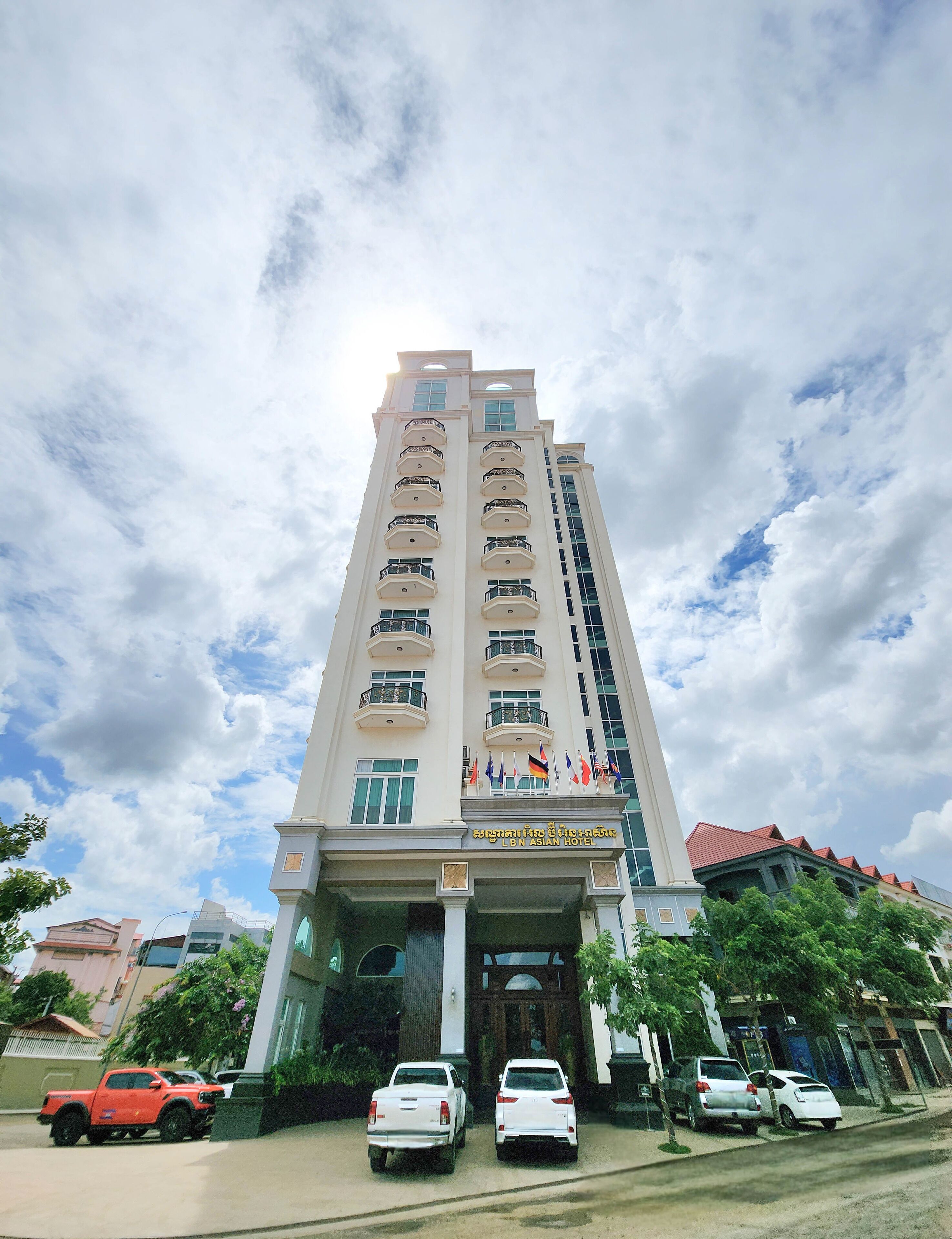 A tall hotel with multiple balconies and flags, featuring a prominent entrance and several parked cars in front.