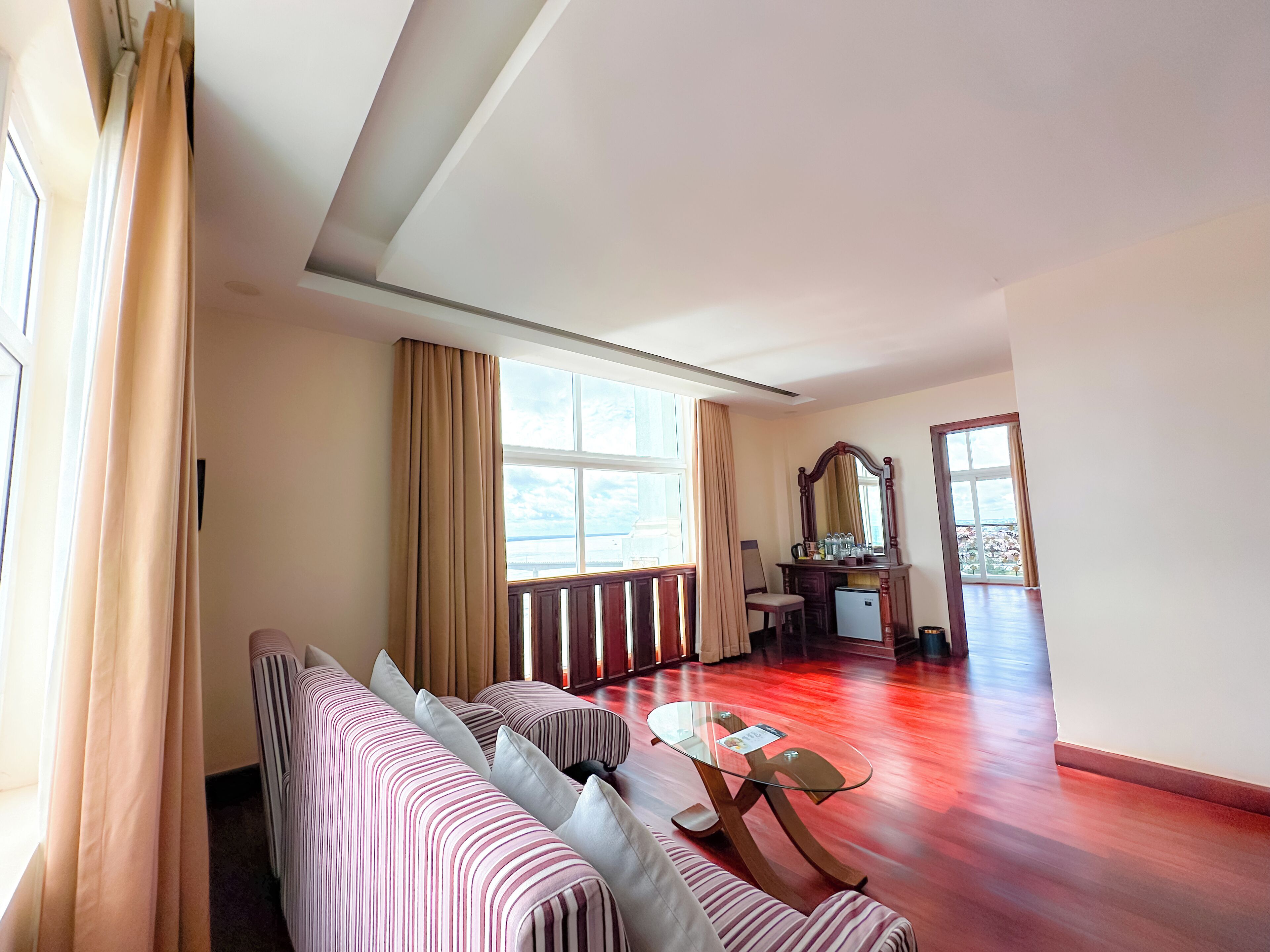 Een hotelkamer met een gestreepte bank, een glazen salontafel, een houten dressoir en een groot raam met gordijnen.