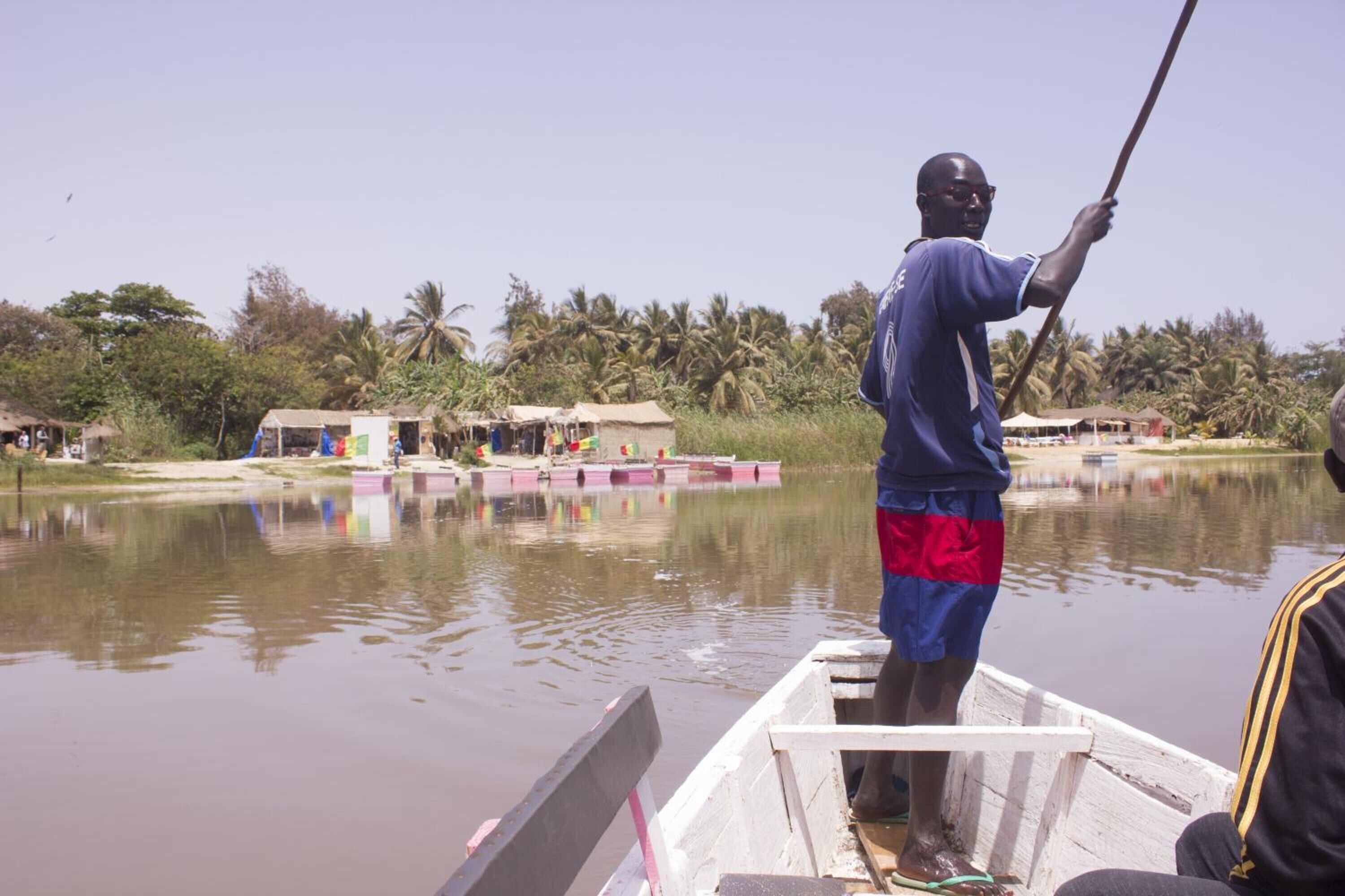 A man standing on a boat in a calm river with houses and palm trees in the background.