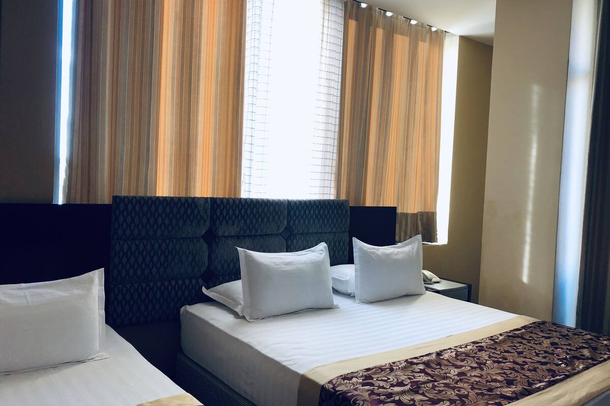 Une chambre d’hôtel avec deux lits, une grande fenêtre avec des rideaux, une tête de lit et une table de chevet avec une lampe.