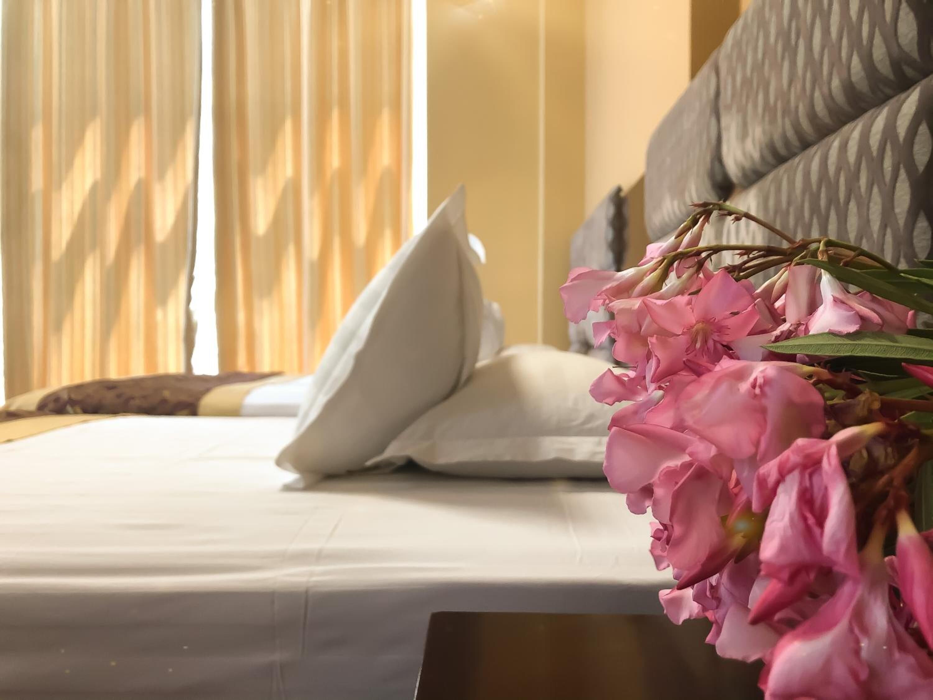 Una cama bien hecha con sábanas y almohadas blancas, un ramo de flores rosas y una ventana con cortinas traslúcidas.