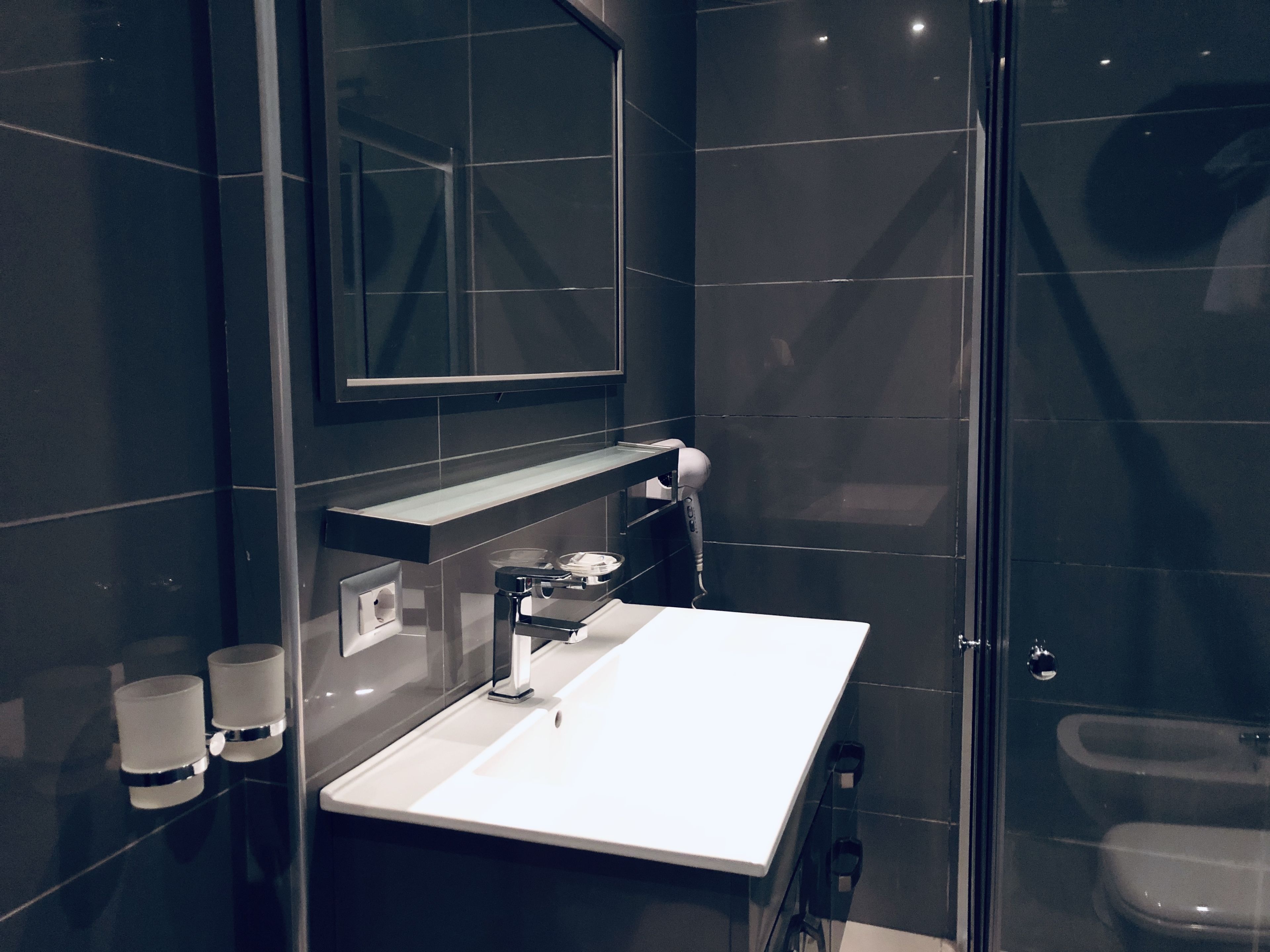 A modern bathroom with a white sink, dark tiles, a glass shower enclosure, and a mirror above the sink.