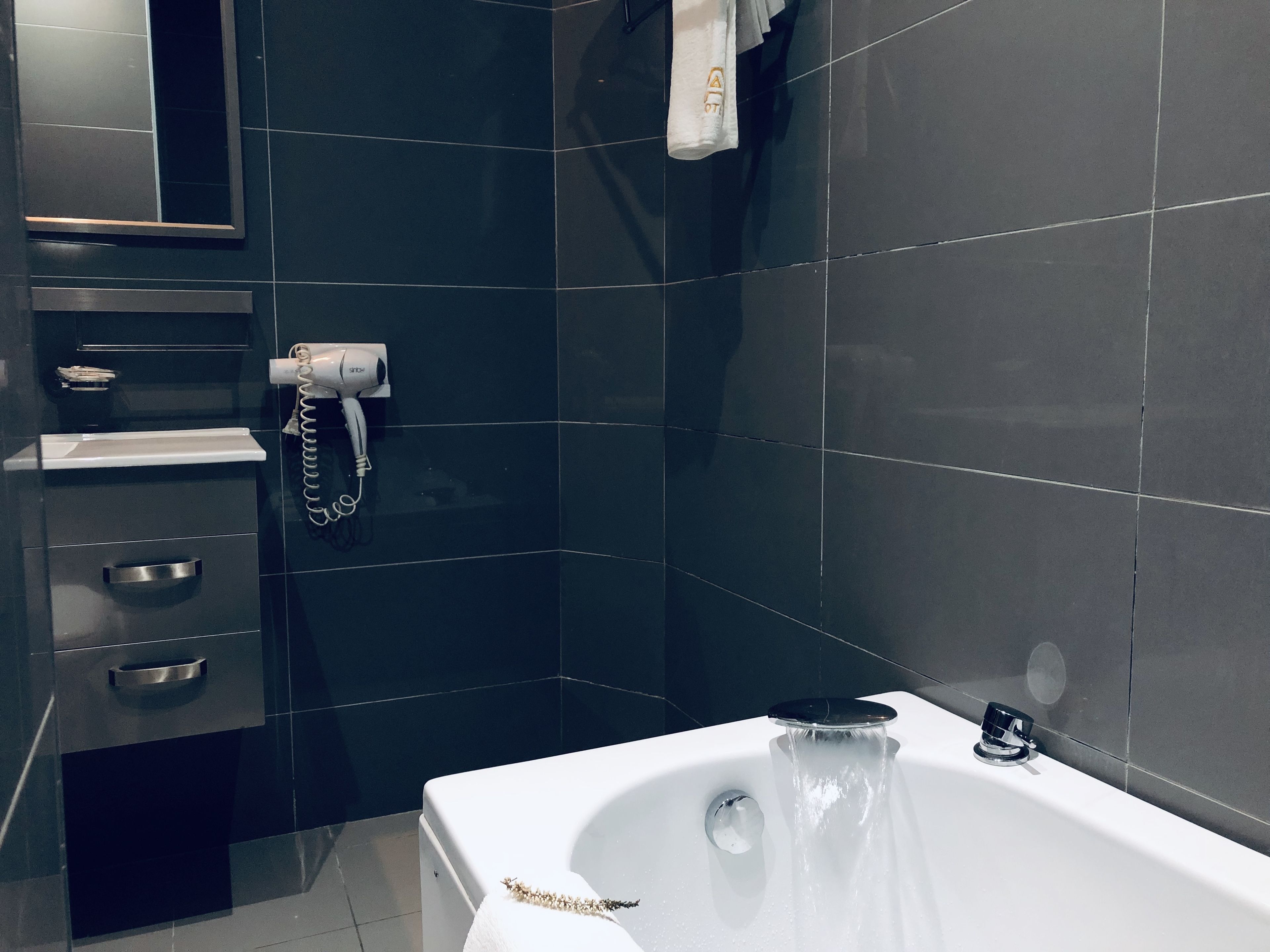 A modern bathroom with dark tiles, a white bathtub, a hairdryer, and a towel rack.