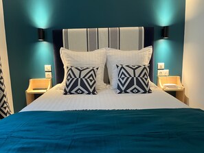 A neatly made bed with a teal bedspread, two patterned pillows, and a striped headboard. There are bedside tables with lamps and a wall socket. - Hotel Chilhar (Espelette)