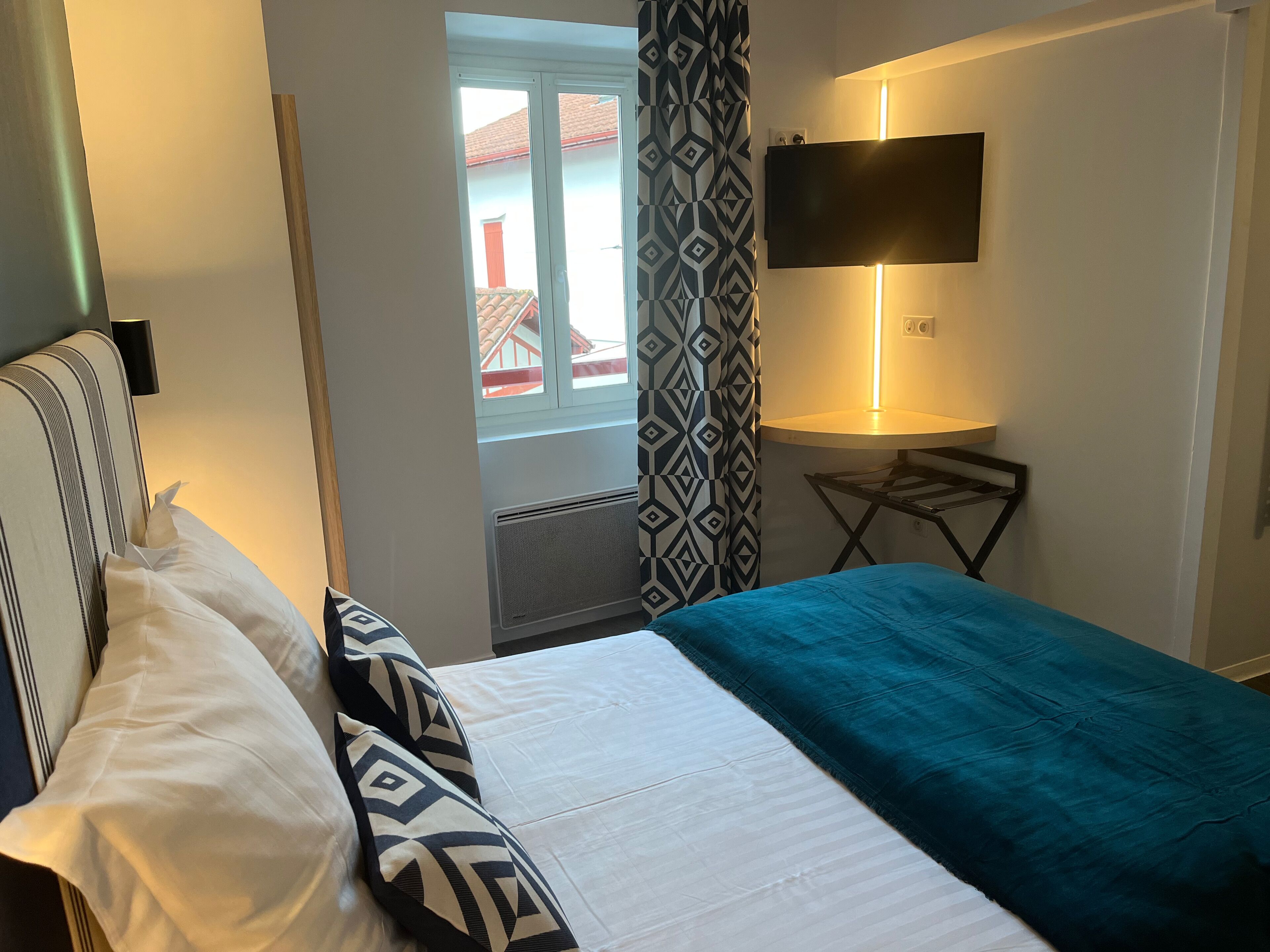 A hotel room with a bed, a TV mounted on the wall, a window with curtains, and a small table with a lamp.