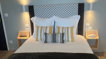Un lit bien fait, avec une tête de lit noire, une literie blanche et des oreillers à rayures. Il y a deux tables de chevet avec des lampes, et un luminaire fixé au mur au-dessus du lit.