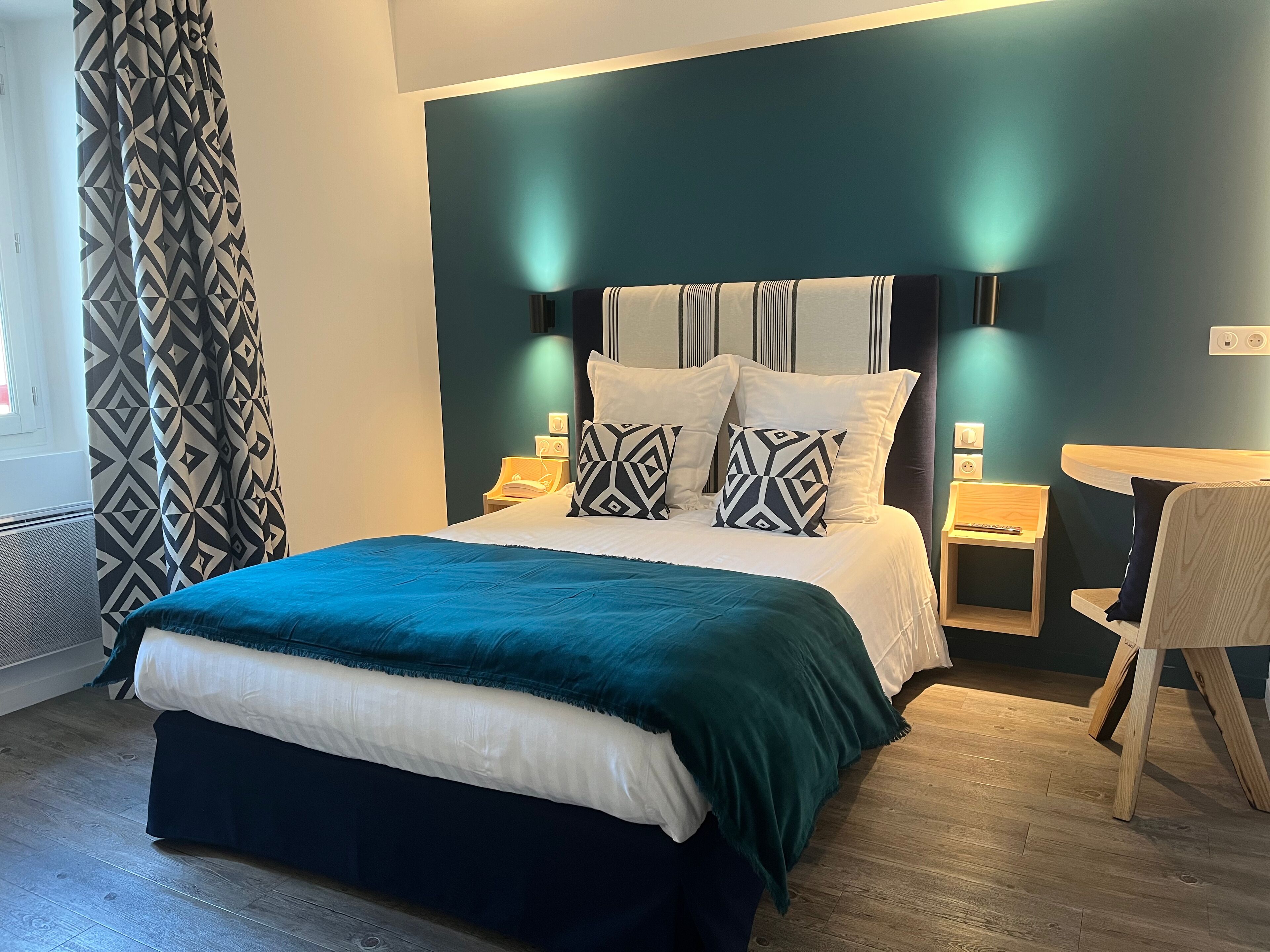 Una cama bien hecha con colcha verde azulada y blanca, dos mesitas de noche con lámparas, una silla y un aplique de pared.