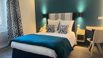 Una cama bien hecha con colcha verde azulada y blanca, dos mesitas de noche con lámparas, una silla y un aplique de pared.