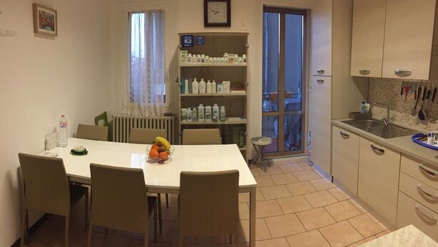 A compact kitchen with a dining area, featuring a white table, chairs, a fruit bowl, and a shelf with various bottles. - Nightstars B&B (Mestre)