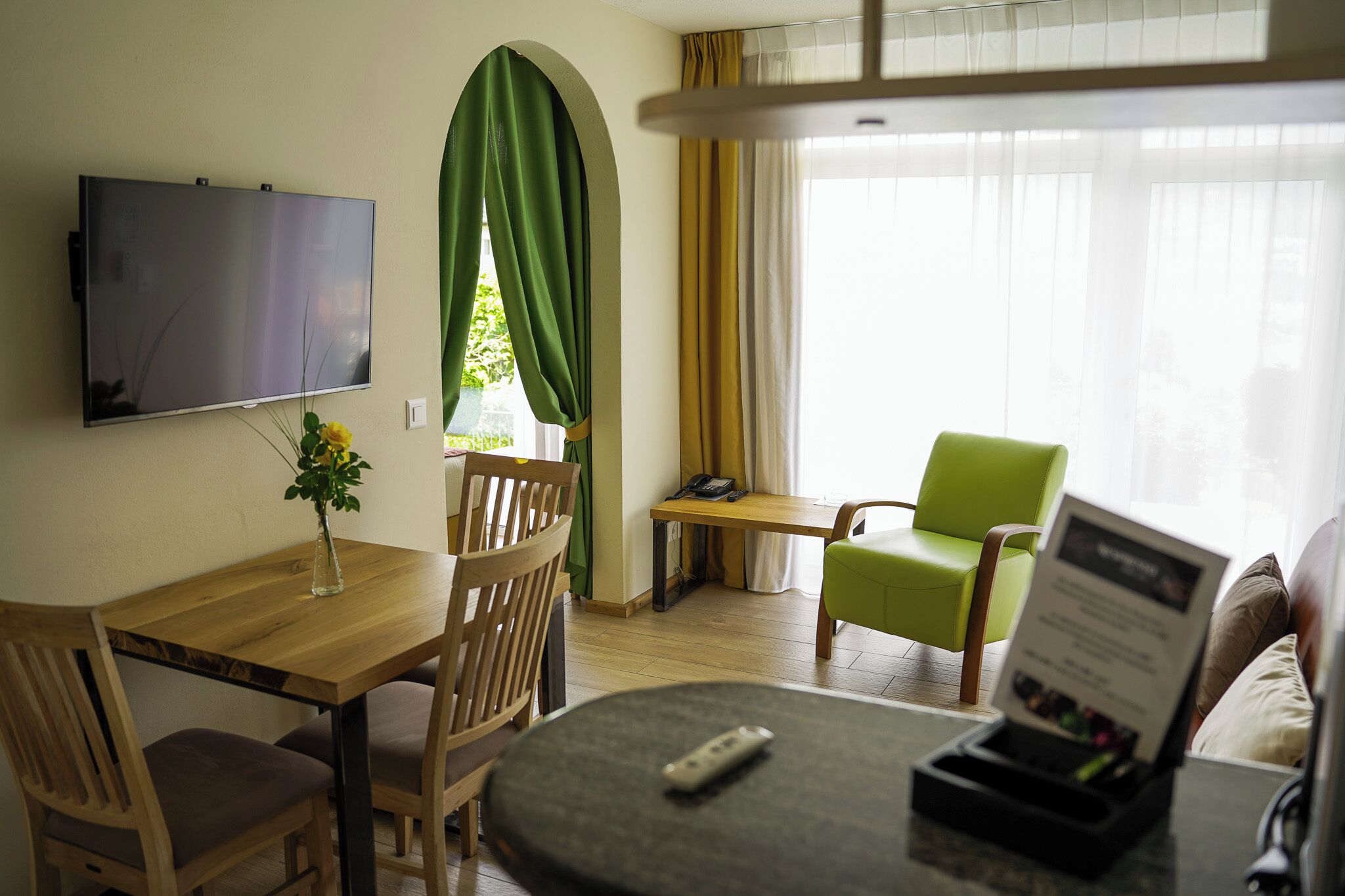 Una habitación con televisión montada en la pared, una mesa de comedor con sillas, una mesita con un teléfono, una silla verde y un sofá.