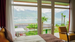A hotel room with a large window offering a view of a coastal town and mountains. - Hotel La Campagnola (Gambarogno)