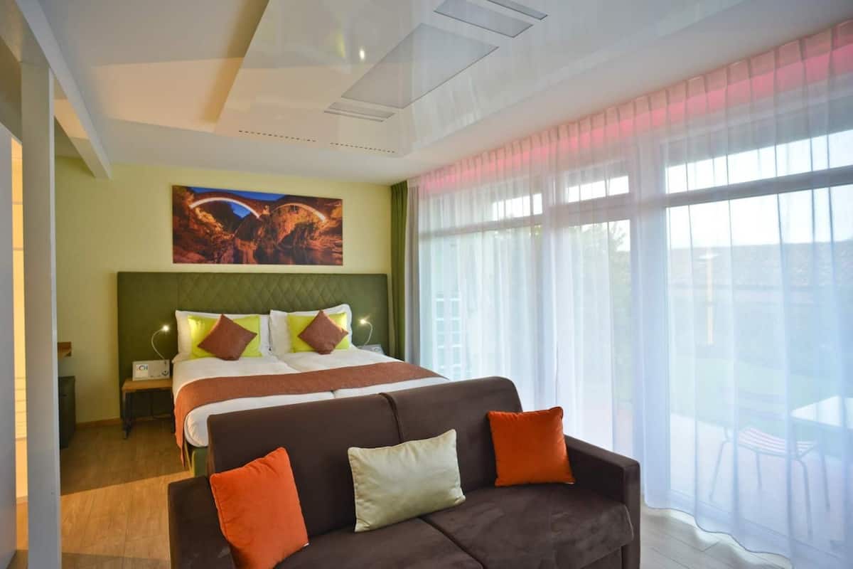 Een hotelkamer met een groot bed, een groen hoofdeinde, een bruine bank en een groot raam met dunne gordijnen.