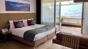 A modern hotel room with a large bed, a nightstand, a desk, and a view of a mountainous landscape through a large window. - Hotel La Campagnola (Gambarogno)