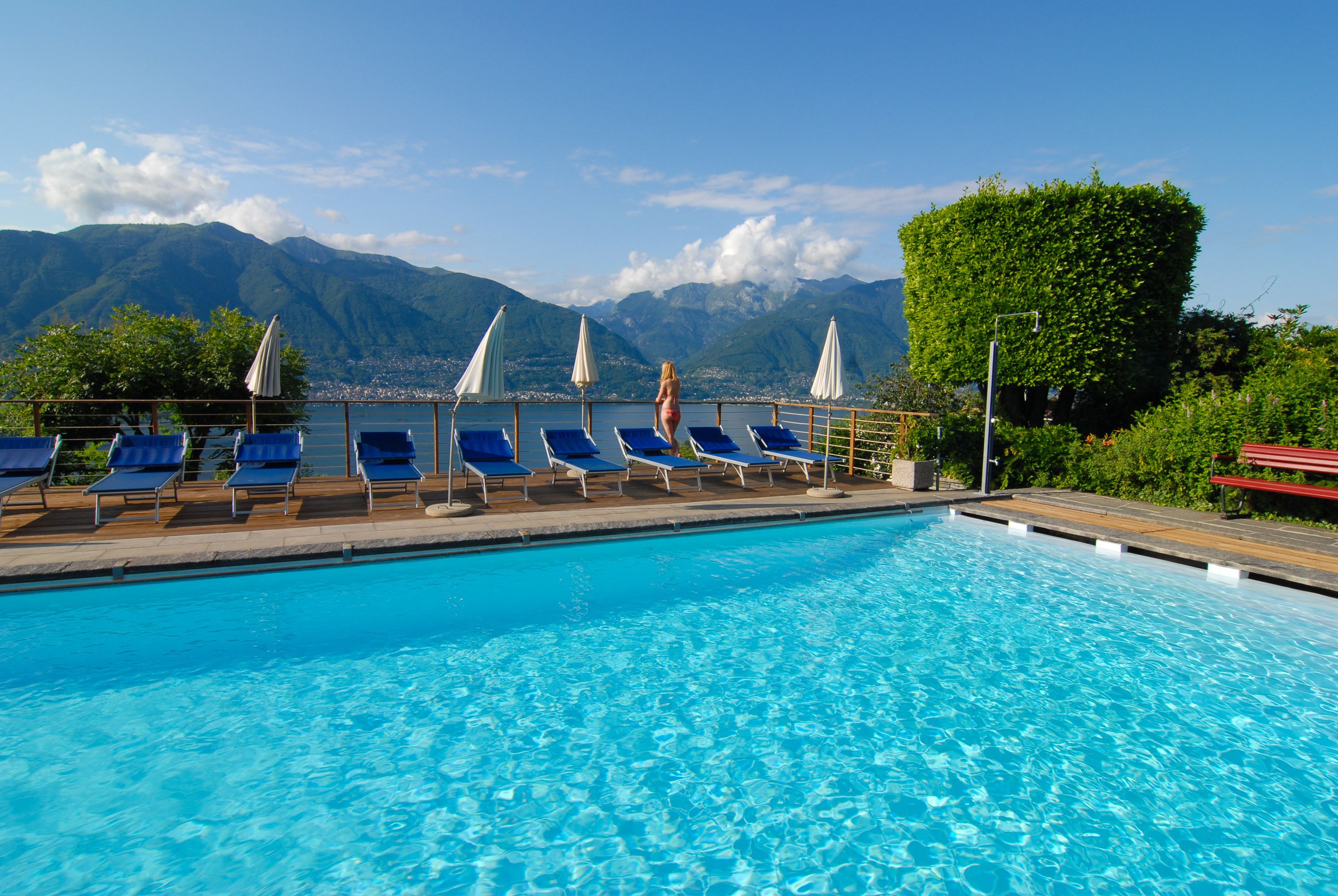 Ein Swimmingpool mit blauen Liegestühlen und Sonnenschirmen, umgeben von Bergen und viel Grün.