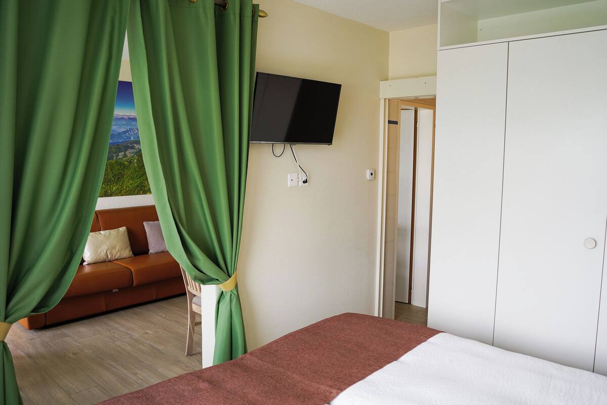 Een kamer met een bed, een aan de muur bevestigde televisie, een bank en uitzicht op een berglandschap door groene gordijnen.